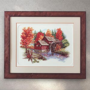 Könnte beinhalten: Gerahmtes Kreuzstich-Kunstwerk, das eine ländliche Herbstszene darstellt. Das Bild zeigt eine rote Wassermühle mit einem Wasserrad, einen Holzzaun und leuchtendes Herbstlaub in Rot-, Orange- und Grüntönen. Der Rahmen ist aus dunkelbraunem Holz.