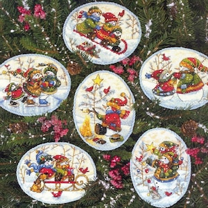 May include: Six embroidered Christmas tree decorations. Each oval decoration depicts a winter scene with snowmen, birds, and trees. The snowmen wear hats and scarves. The decorations are set against a backdrop of evergreen branches.