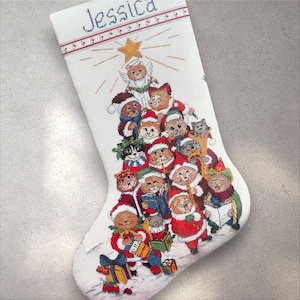 May include: White Christmas stocking with a cross-stitch design featuring a tree made of cats wearing Santa hats and holding gifts. The name "Jessica" is stitched at the top, with a star and angel cat at the top.