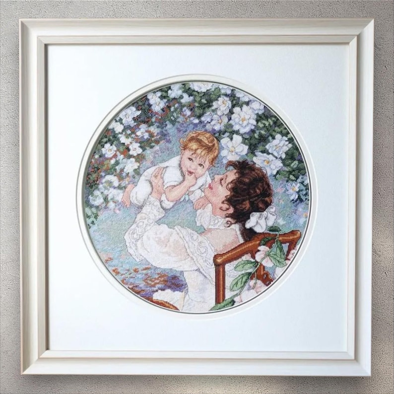 May include: Framed artwork featuring a mother and child in a garden setting. The artwork is a cross-stitch design, with the mother holding the child. The artwork is framed in a white frame.