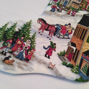 May include: Christmas stocking with a detailed tapestry scene of a snowy village. The design features figures in winter attire, horse-drawn sleighs, and decorated buildings. The color palette includes reds, greens, and creams.