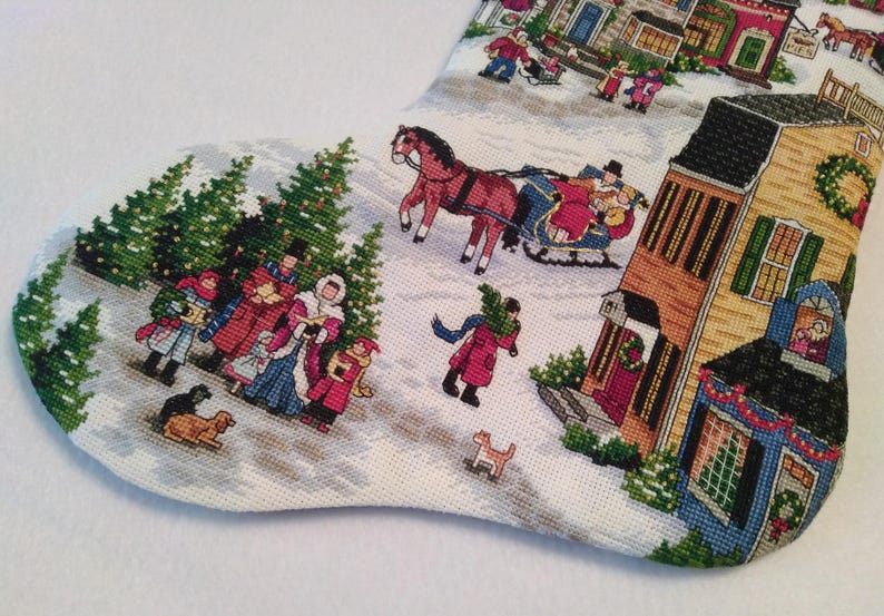 May include: Christmas stocking with a detailed tapestry scene of a snowy village. The design features figures in winter attire, horse-drawn sleighs, and decorated buildings. The color palette includes reds, greens, and creams.