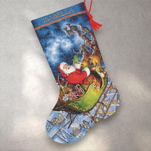 May include: A Christmas stocking featuring Santa Claus in his sleigh, pulled by reindeer, against a night sky backdrop. The stocking is blue with the name "ISABELLA" at the top and a red tassel. The bottom of the stocking depicts a snowy village.