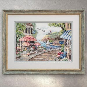 Könnte beinhalten: Gerahmtes Kreuzstich-Kunstwerk, das eine Küstenstadt zeigt. Das Bild zeigt ein Café mit rot-weiß gestreiften Markisen, einen Leuchtturm und ein Segelboot auf dem Wasser. Der Rahmen ist hellblau.