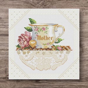Puede incluir: Diseño de punto de cruz con una taza de té con la palabra "Mother" y detalles florales. Se incluye una rosa rosa y otros elementos florales. El diseño está sobre un fondo blanco con un borde decorativo.