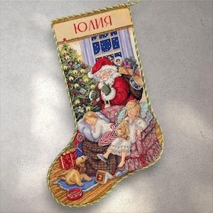 May include: Christmas stocking depicting Santa Claus, children, and a dog. The beige stocking has a green trim and the name "Юлия" in red. The scene includes a Christmas tree and a snowy window.