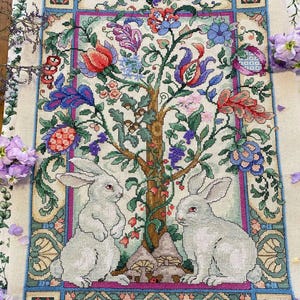 May include: A detailed cross-stitch tapestry featuring two white rabbits beneath a stylized tree with colorful flowers and foliage. The design includes a border with floral and geometric patterns, showcasing a blend of green, blue, pink, and red hues.