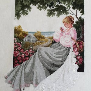 May include: Cross-stitch artwork depicting a woman in a pink dress seated outdoors. The scene includes a house, flowers, and a tree, with a coastal view in the background. The needlework is detailed, showcasing a serene, pastoral theme.