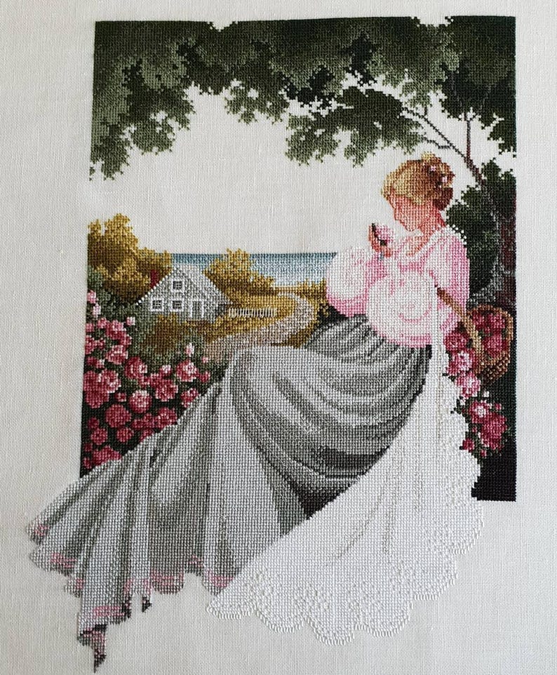 May include: Cross-stitch artwork depicting a woman in a pink dress seated outdoors. The scene includes a house, flowers, and a tree, with a coastal view in the background. The needlework is detailed, showcasing a serene, pastoral theme.
