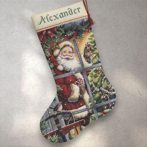 Könnte beinhalten: Roter Weihnachtsstrumpf mit einer Illustration von Santa Claus, der durch ein Fenster blickt. Der Strumpf zeigt auch einen Vogel und einen Weihnachtsbaum. Der Name "Alexander" ist oben eingestickt.