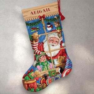 Puede incluir: Un calcetín navideño con un diseño festivo que presenta a Papá Noel, un niño y varios juguetes. El nombre "ABIGAIL" se muestra en la parte superior. El calcetín está decorado con una escena de ventana y una borla roja.