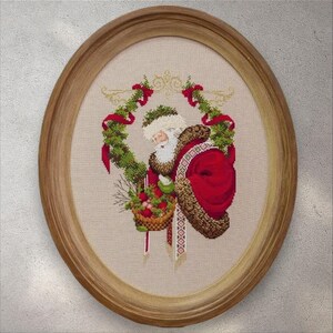 May include: Oval framed cross-stitch of Santa Claus, wearing a red suit with brown fur trim, holding a basket of fruit. He is surrounded by a green wreath with red ribbons and gold accents. The frame is made of light brown wood.