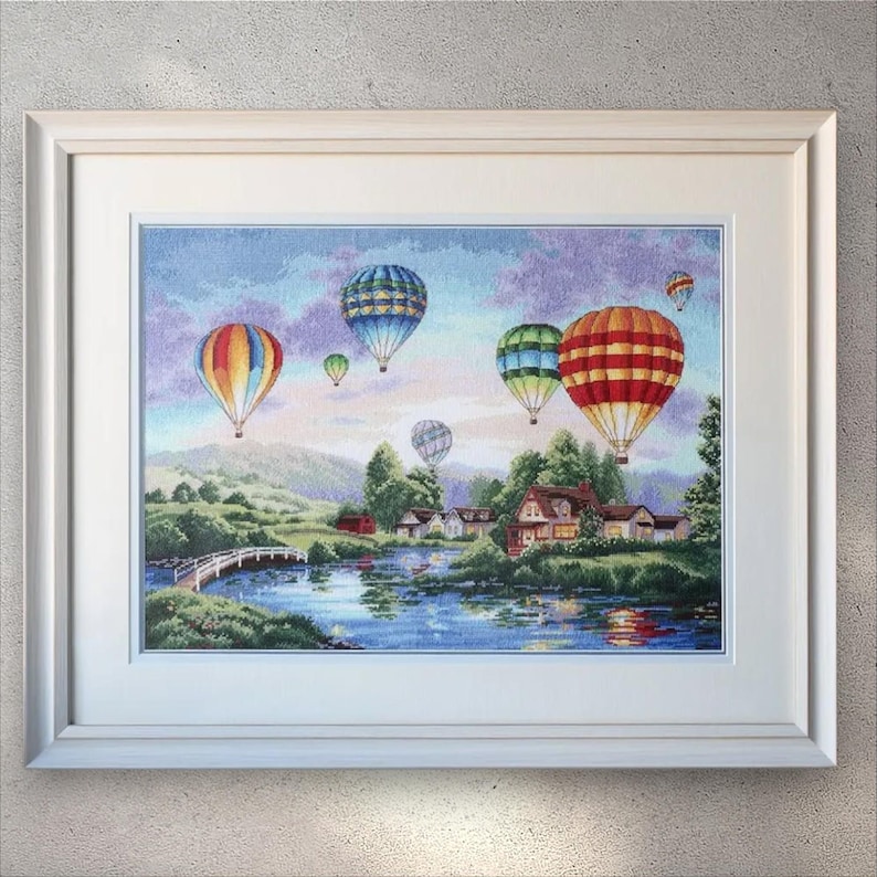 Puede incluir: Arte en punto de cruz enmarcado que representa un paisaje sereno con globos aerost&aacute;ticos de colores flotando sobre un lago y casas. La obra de arte presenta un marco blanco y una escena detallada y colorida.
