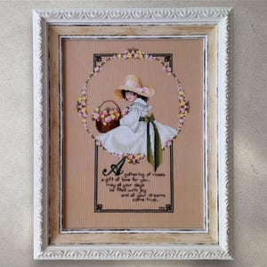 May include: Cross-stitch artwork in a white ornate frame. The image features a girl in a white dress and hat, holding a basket of roses. A floral wreath surrounds the girl. Text at the bottom reads "A gathering of roses, a gift of love for you, may all your days be filled with joy and all your dreams come true."