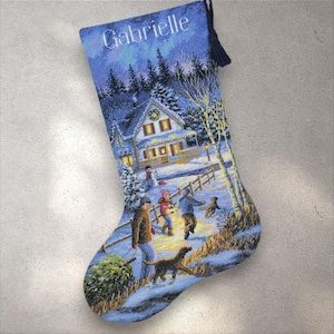 May include: Blue Christmas stocking with a winter scene featuring a house, people, and dogs. The word "Gabrielle" is embroidered at the top. The stocking has a tassel and is made of fabric.