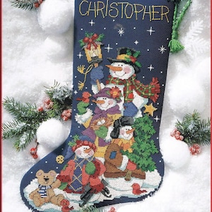 May include: A navy blue Christmas stocking with the name "CHRISTOPHER" embroidered in gold. The stocking features a cross-stitch design of snowmen, a teddy bear, and a Christmas tree, all set against a snowy background.