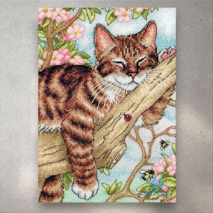 Op de afbeelding: Kruissteekkunstwerk van een lapjeskat die op een tak slaapt. De kat heeft bruine en bruine strepen, gesloten ogen en lange witte snorharen. Roze bloemen en groene bladeren omringen de kat, met een lieveheersbeestje en bijen zichtbaar. De achtergrond is lichtblauw.