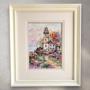 May include: Framed cross-stitch art of a lighthouse on a rocky cliff, with a red and white striped tower. The scene includes a small house, a path, and colorful flowers. The artwork is set against a sunset sky and framed in a white wooden frame.