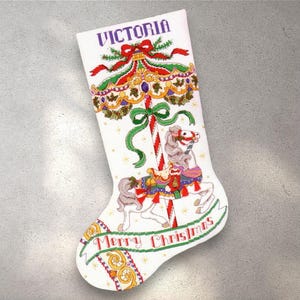 May include: White Christmas stocking with a carousel horse design. The stocking features the name "Victoria" in purple, a red and green bow, and the words "Merry Christmas". The carousel horse is gray and white with colorful accents.