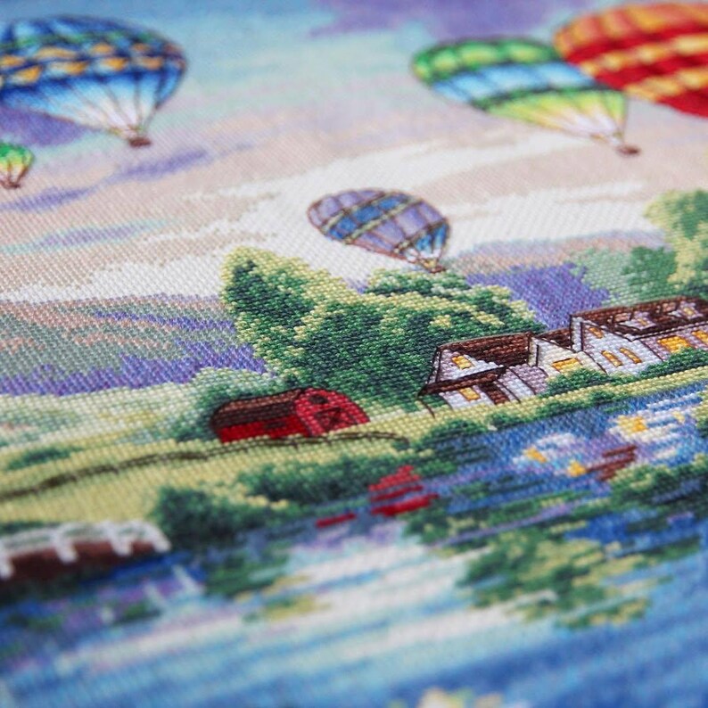 Puede incluir: Un paisaje bordado a punto de cruz con globos aerost&aacute;ticos de colores flotando en el cielo. La escena incluye un granero rojo, casas y una masa de agua reflectante, todo ello realizado con puntadas detalladas. El cielo cambia de azul a morado.