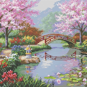 May include: A cross-stitch landscape depicts a serene Japanese garden scene. A red arched bridge spans a river, with cherry blossom trees in full bloom. The scene includes a stone lantern, water lilies, and lush greenery.