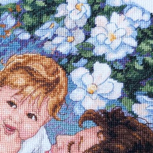 May include: A cross-stitch embroidery featuring two figures with light brown and dark brown hair, surrounded by white and light blue flowers with yellow centers. The background is a mix of green and blue.
