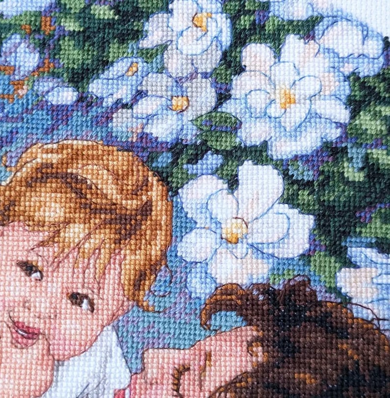 May include: A cross-stitch embroidery featuring two figures with light brown and dark brown hair, surrounded by white and light blue flowers with yellow centers. The background is a mix of green and blue.