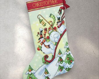 May include: A festive Christmas stocking with a light green backdrop, decorated with snowmen, Christmas trees, and the name "CHRISTOPHER" in red. It has a red trim and a red hanging loop.