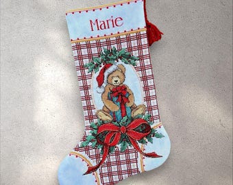 May include: Christmas stocking featuring a teddy bear in a Santa hat, holding a present. The stocking has a red and white plaid design, with the name "Marie" embroidered at the top. A red tassel is attached.