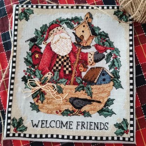 May include: Cross-stitched Christmas decoration featuring Santa Claus surrounded by birdhouses and birds, with holly and the text "WELCOME FRIENDS." The design is framed with a black and white border and a beige background.