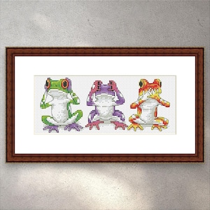 Könnte beinhalten: Gerahmte Kreuzsticharbeit mit drei bunten Fröschen. Jeder Frosch, in Grün, Lila und Orange, bedeckt Augen, Ohren und Mund. Das Kunstwerk befindet sich vor einem weißen Hintergrund, in einem braunen Holzrahmen.
