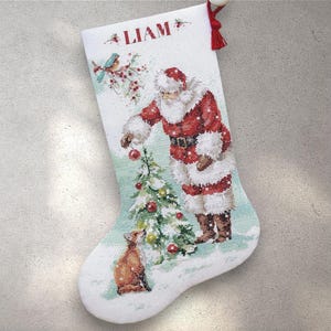 May include: White Christmas stocking with a festive illustration of Santa Claus decorating a Christmas tree. The stocking features the name "LIAM" in red letters and a small fox. A red tassel hangs from the top.