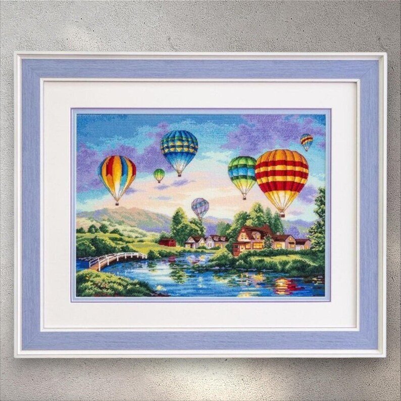 Puede incluir: Obra de arte en punto de cruz enmarcada que representa un paisaje con globos aerost&aacute;ticos de colores sobre un r&iacute;o y casas. La obra est&aacute; enmarcada en un marco de madera azul claro con un paspart&uacute; blanco.