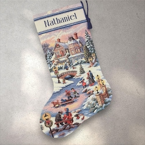 Pode incluir: Uma meia de Natal com uma cena de inverno com uma ponte, casas e pessoas. A meia é decorada com uma paisagem de inverno e o nome "Nathaniel" é exibido no topo.