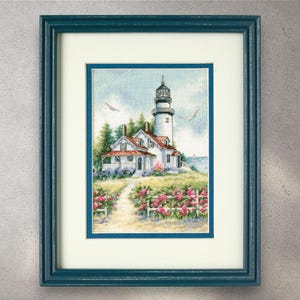 May include: Framed cross-stitch art of a lighthouse and adjacent house on a grassy hill, with a path leading to the buildings. The artwork features a blue sky, white fence, and pink flowers. The frame is teal.