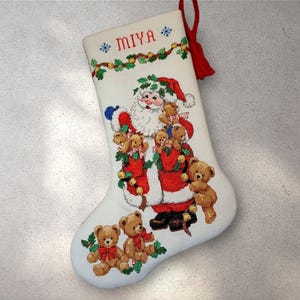 May include: White Christmas stocking with a cross-stitch design featuring Santa Claus, teddy bears, and holly. The name "MIYA" is embroidered at the top. A red tassel hangs from the top. Festive holiday decor.