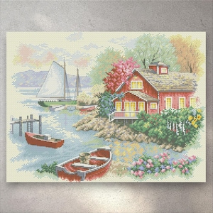 May include: Cross-stitch artwork showing a coastal scene. A red house with yellow windows sits on a rocky shore, surrounded by greenery and flowers. Two red rowboats float in the water near a small pier. Sailboats are in the distance.
