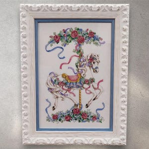 May include: Cross-stitch picture of a carousel horse decorated with roses and ribbons. The horse is white with a pink and blue saddle, surrounded by floral details. The artwork is framed in a white, ornate frame with a blue inner border.