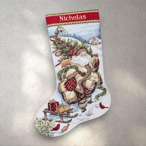 Pode incluir: Meia de Natal com uma cena de inverno com o Pai Natal, uma árvore de Natal e um trenó com presentes. A parte superior da meia é vermelha com o nome "Nicholas" bordado em branco.