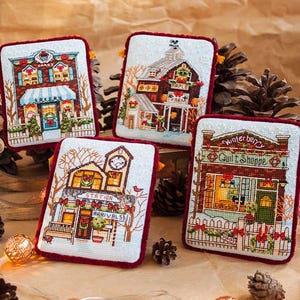 Peut inclure: Quatre pièces décoratives brodées avec une bordure et des glands rouges. Chaque pièce représente un bâtiment différent : une boulangerie, une ferme, une gare et une boutique de courtepointes. Les bâtiments sont détaillés au point de croix, avec des décorations de vacances festives et des scènes d'hiver.