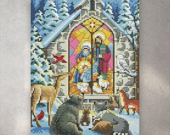 May include: Cross-stitch artwork depicting a Nativity scene inside a church with a stained-glass window. The artwork features a variety of animals, including deer, a bear, and a raccoon, in a winter landscape with snow-covered trees.