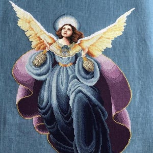 May include: A cross-stitch embroidery of an angel with golden wings, a blue dress, and a purple robe. The angel has a halo and is looking upwards.