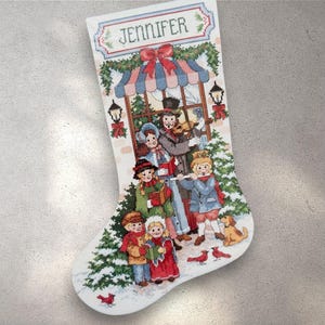 Può includere: Calza di Natale bianca con un disegno a punto croce. La calza presenta una scena festosa con cantori, un cane e il nome "JENNIFER" in alto. Il design include accenti rossi, verdi e blu, creando un tema festivo.