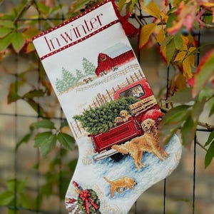 May include: A Christmas stocking with a winter scene. The stocking features a red truck carrying a Christmas tree, golden retrievers, and the text "In Winter". The background includes a red barn and snowy landscape.