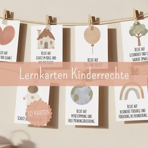 Puede incluir: Un conjunto de diez tarjetas ilustradas sobre los derechos de los niños, colgadas de una cuerda con pinzas de madera. Cada tarjeta presenta una ilustración sencilla y texto en alemán, incluyendo un corazón, una casa, un globo, un árbol, un astronauta, un globo terráqueo y un arcoíris.