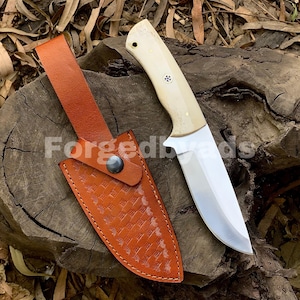 May include: A hunting knife with a cream-colored handle and a polished steel blade rests beside a brown leather sheath. The sheath has a basketweave pattern and a snap closure. The text "ForgedByAds" is visible.