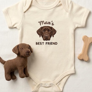 Könnte beinhalten: Cremefarbener Baby-Body mit einer braunen Hundeillustration und dem Text "Max's BEST FRIEND". Ein braunes Stofftier in Hundeform und ein Holzknochen-Spielzeug liegen daneben.