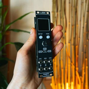 May include: A black electronic device with a small screen and multiple input/output ports. The device has buttons labeled "SETUP" and "MODE", and the text "TEMBRA NAKIMIA" is visible. The device is held in a hand.