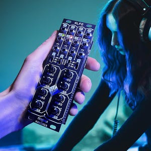 Puede incluir: Un módulo de sintetizador de música electrónica negro con la etiqueta "4LFO" se sostiene en una mano. El módulo tiene numerosas perillas y puertos. Una persona con auriculares y un atuendo negro está operando un sintetizador más grande. La escena está iluminada con tonos azules y verdes.