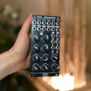 May include: A black Dual Sequencer module with numerous knobs, jacks, and the text "DUAL SEQUENCER." The module has the text "TEMBRA KAKIMIA" at the bottom. The module is held in a hand.